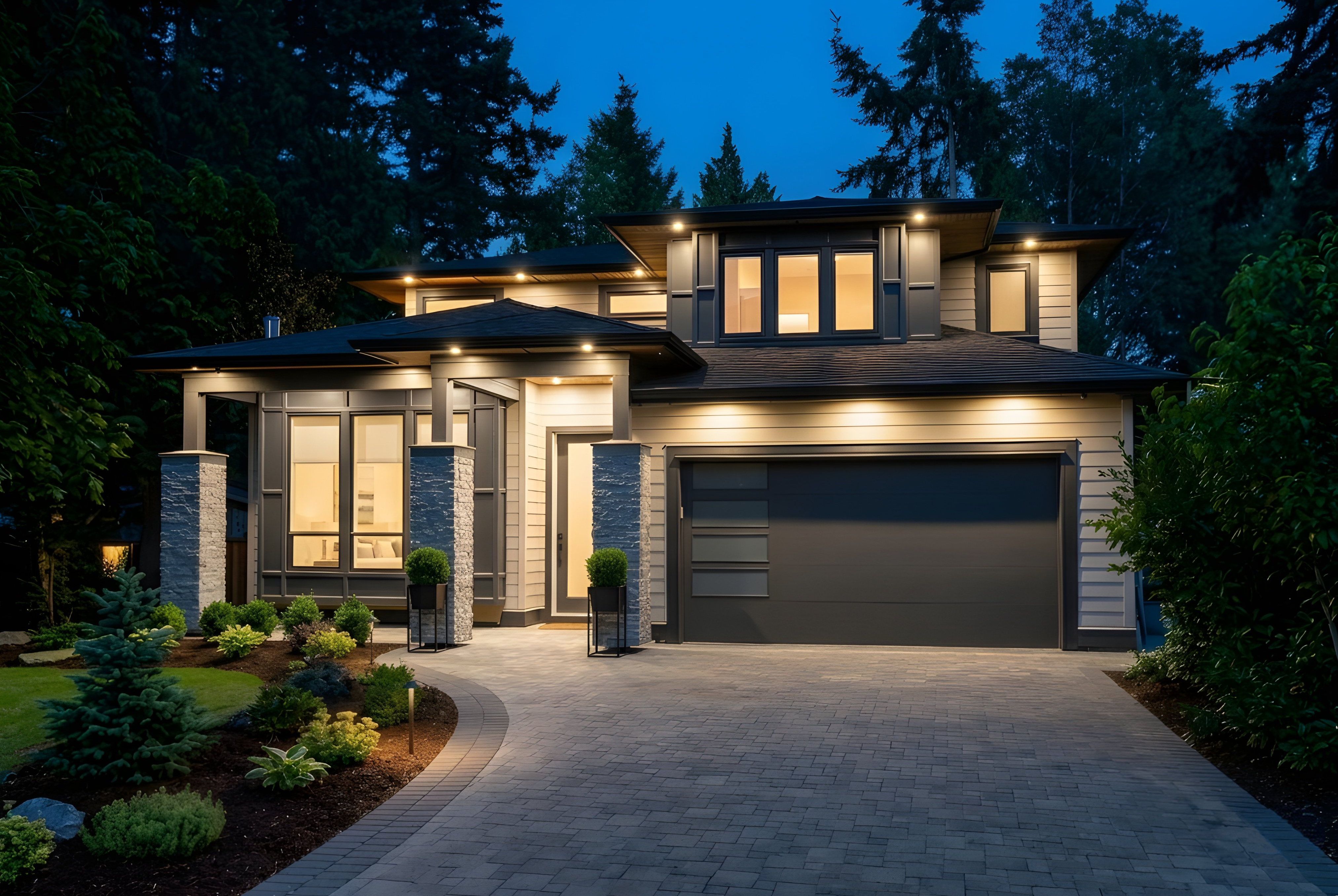 Modern two-storey home at twilight with warm interior lights, stone-column entry and paver driveway