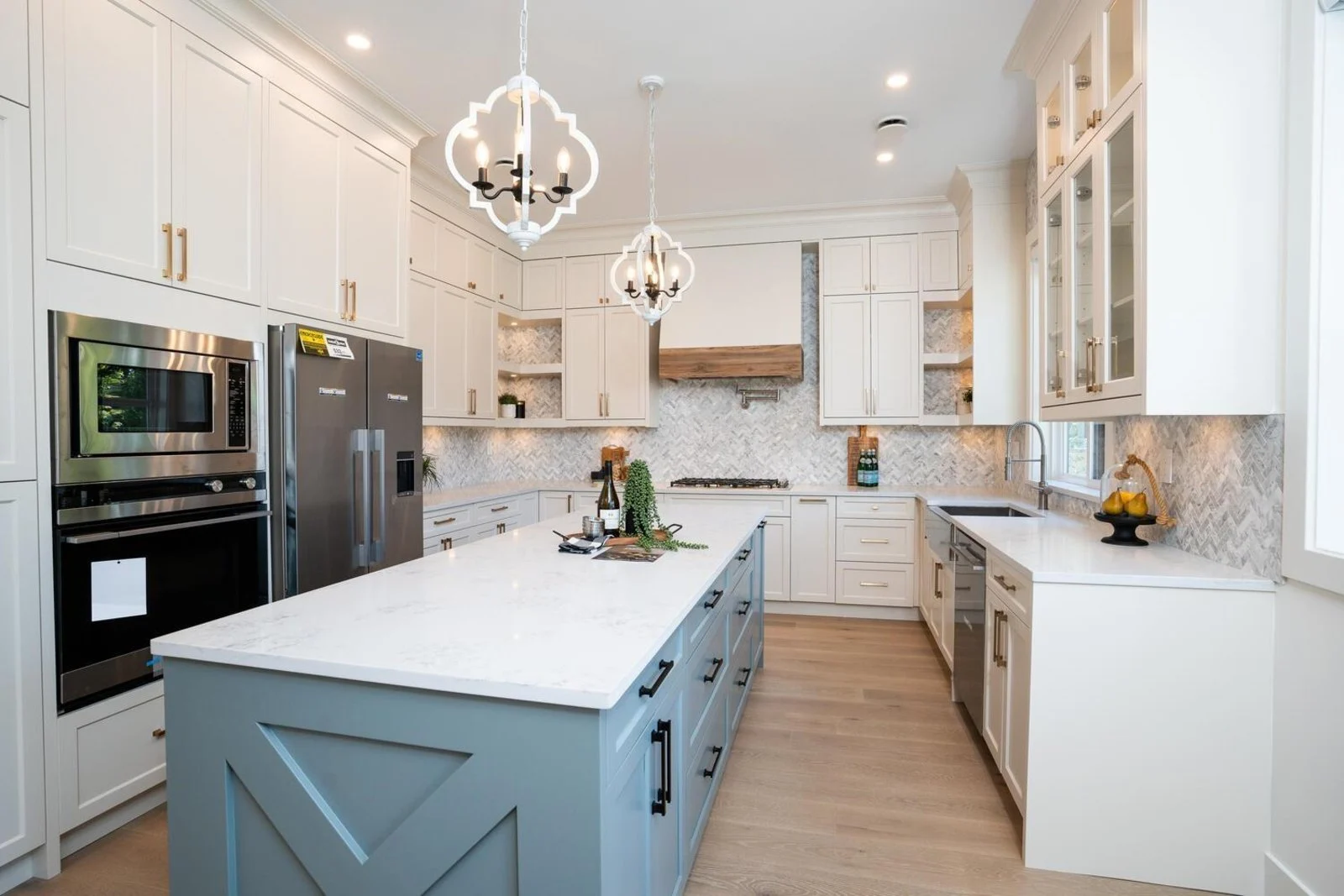 Sage-blue kitchen island with paired pendant chandeliers and marble herringbone backsplash