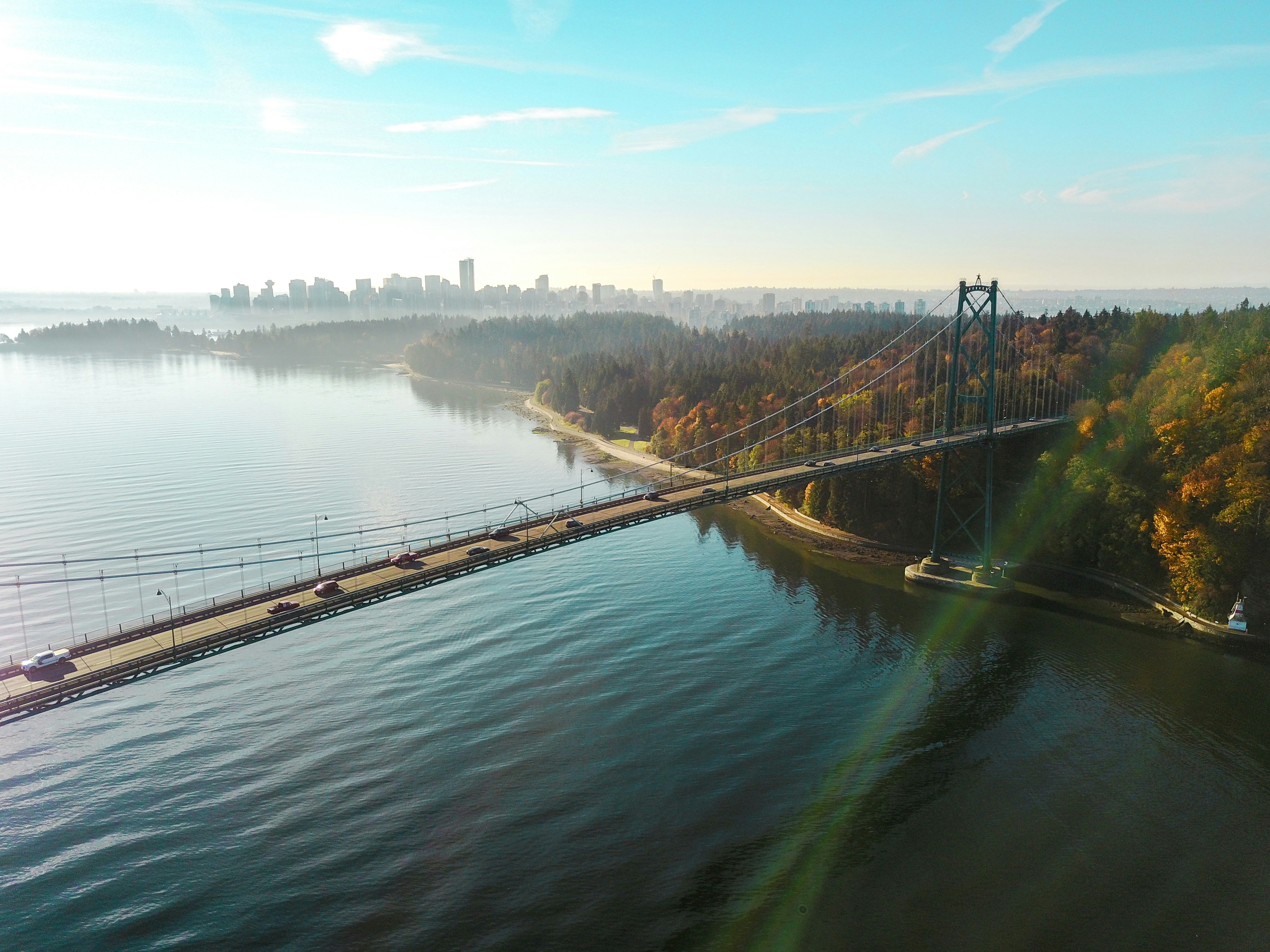 Aerial view of the Lions Gate Bridge crossing Burrard Inlet at golden hour, with Stanley Park's autumn forest and the downtown Vancouver skyline in the distance